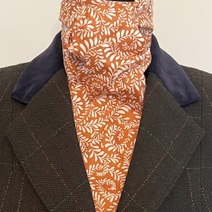 May include: A patterned ascot in shades of orange and white, featuring a botanical design, is worn with a dark brown tweed jacket with a blue collar. The ascot adds a pop of color and pattern to the outfit.