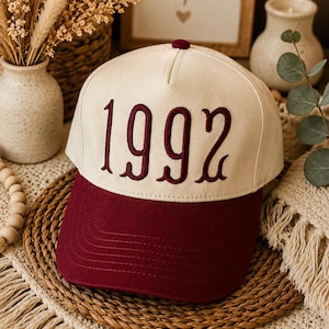 May include: A baseball cap with a cream-colored crown and a dark maroon brim. The front of the crown features the embroidered text 1992 in a dark maroon, stylized font. The cap has a button at the top center and structured panels.