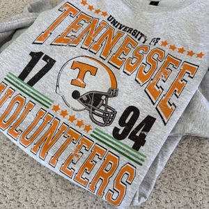 May include: A gray sweatshirt featuring the text "UNIVERSITY OF TENNESSEE VOLUNTEERS" in orange and black. The design includes a football helmet graphic and the numbers "17" and "94". The sweatshirt has a crew neck.