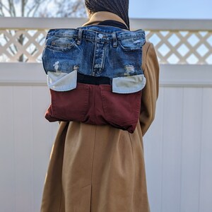 May include: A denim backpack constructed from repurposed jeans. The top section features blue denim with distressed details, while the bottom section is made from burgundy-colored denim. The backpack is worn over a tan coat.