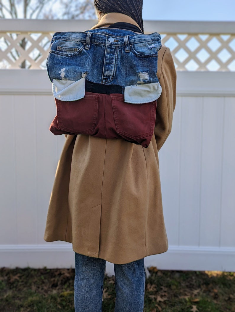 May include: A backpack made from recycled denim, with a patchwork design. The top section is blue denim with distressed areas, and the bottom section is burgundy. The backpack is worn over a tan coat.