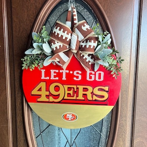 May include: A red and gold round door hanger with the text "Let's Go 49ers" and a football-themed bow. The 49ers logo is in the centre of the gold section.