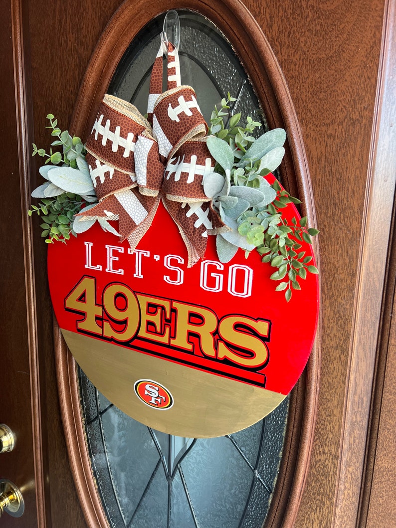 May include: A red and gold round door hanger with the words "Let's Go 49ers" and a football-themed bow. The door hanger has a small San Francisco 49ers logo in the lower right corner.