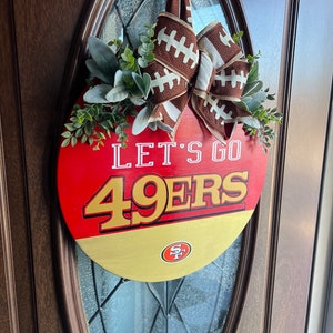 May include: A round door hanger in red, white and gold with the words "Let's Go 49ers" and a football-themed bow. The door hanger features a San Francisco 49ers logo.