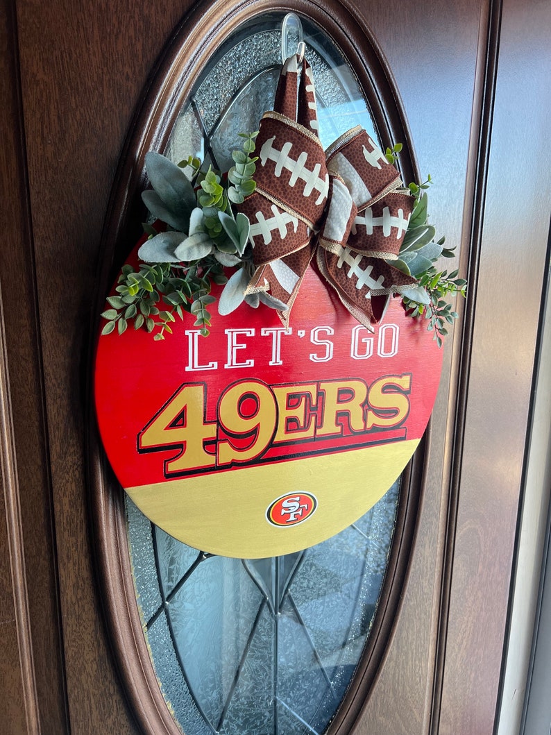 May include: A round door hanger in red, white and gold with the words "Let's Go 49ers" and a football-themed bow. The door hanger features a San Francisco 49ers logo.