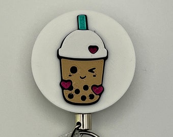 May include: White retractable badge reel with a cartoon bubble tea cup with a winking face and hearts. The bubble tea cup is brown with white milk foam and has a green lid and straw.
