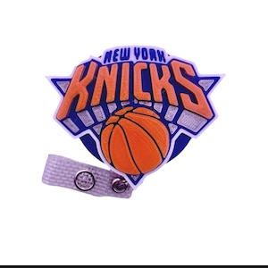 May include: A New York Knicks basketball team logo badge reel featuring the orange and blue team emblem with the text NEW YORK KNICKS above an orange basketball graphic, attached to a white textured fabric strap with a silver metal snap button.