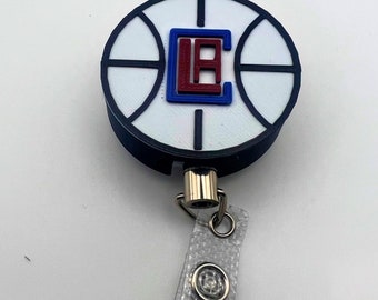May include: A white basketball-shaped retractable badge holder with a black outline and a red and blue "LA" logo. The holder is attached to a white retractable badge reel.