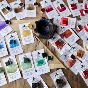 May include: An assortment of watercolour paint pans in various colours, including teal, green, red, gold, and black. The pans are arranged on a wooden surface with their corresponding labels, which feature the brand name "Earth & Essence essee" and the colour name.