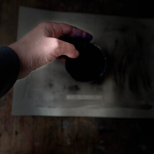 Op de afbeelding: Een hand houdt een klein, rond, zwart bakje vast met een donkerblauwe vloeistof erin. Het bakje staat op een wit oppervlak.