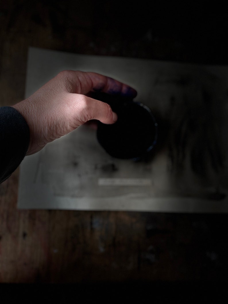 Op de afbeelding: Een hand houdt een klein, rond, zwart bakje vast met een donkerblauwe vloeistof erin. Het bakje staat op een wit oppervlak.