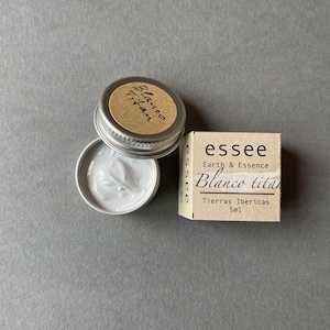 Pu&ograve; includere: Un piccolo barattolo d'argento con una crema bianca al suo interno. Il barattolo ha un'etichetta marrone che dice "Blanco Titan" e "Tierras Ibericas 5ml". L'etichetta ha anche il testo "essee Earth & Essence".