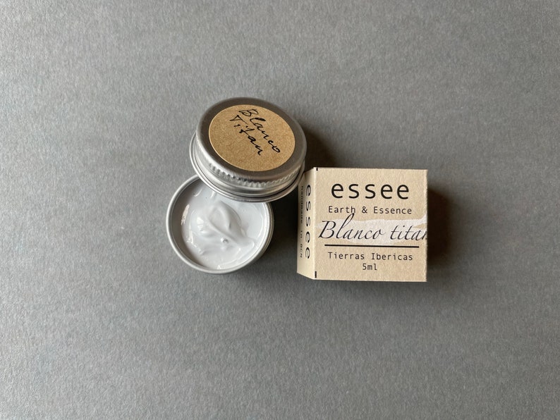 Pu&ograve; includere: Un piccolo barattolo d'argento con una crema bianca al suo interno. Il barattolo ha un'etichetta marrone che dice "Blanco Titan" e "Tierras Ibericas 5ml". L'etichetta ha anche il testo "essee Earth & Essence".
