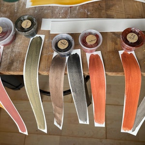 Puede incluir: Siete peque&ntilde;os recipientes de pintura de varios colores, incluyendo dorado, marr&oacute;n, negro, rojo y naranja. Cada recipiente est&aacute; etiquetado con un nombre y se encuentra sobre una tira de papel blanco con una muestra pintada del color.