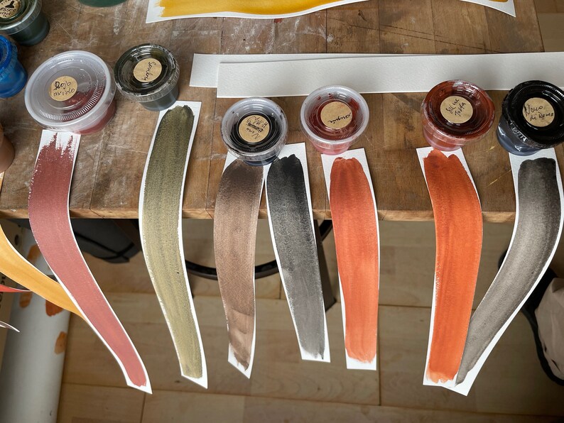 Puede incluir: Siete peque&ntilde;os recipientes de pintura de varios colores, incluyendo dorado, marr&oacute;n, negro, rojo y naranja. Cada recipiente est&aacute; etiquetado con un nombre y se encuentra sobre una tira de papel blanco con una muestra pintada del color.