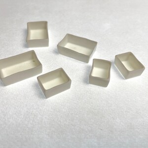 May include: A collection of six small, rectangular, open-top containers. The containers have a light beige exterior and a white interior. They are arranged on a white surface.