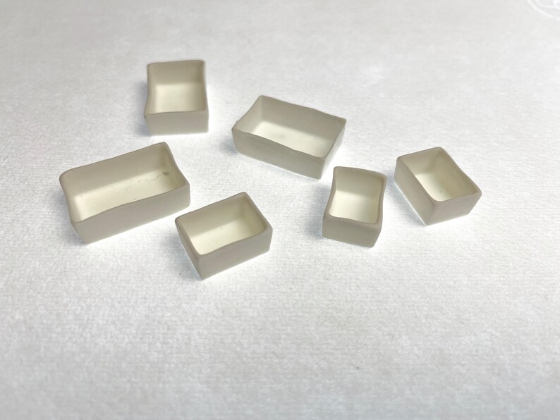 May include: A collection of six small, rectangular, open-top containers. The containers have a light beige exterior and a white interior. They are arranged on a white surface.