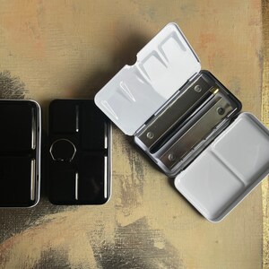May include: A set of three black and white watercolor paint palettes. One palette is open, revealing two metal mixing trays and a white interior. The other two palettes are closed, showing a glossy black exterior. The palettes are on a textured, neutral-toned surface.