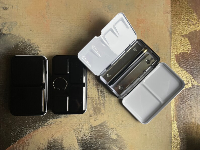 May include: A set of three black and white watercolor paint palettes. One palette is open, revealing two metal mixing trays and a white interior. The other two palettes are closed, showing a glossy black exterior. The palettes are on a textured, neutral-toned surface.