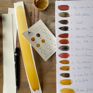 Puede incluir: Una carta de pintura acuarela con 14 tonos diferentes de marr&oacute;n, naranja y amarillo. La carta est&aacute; etiquetada con los nombres italiano e ingl&eacute;s de cada color. La carta est&aacute; en una hoja de papel blanco y est&aacute; rodeada de otros materiales de arte.
