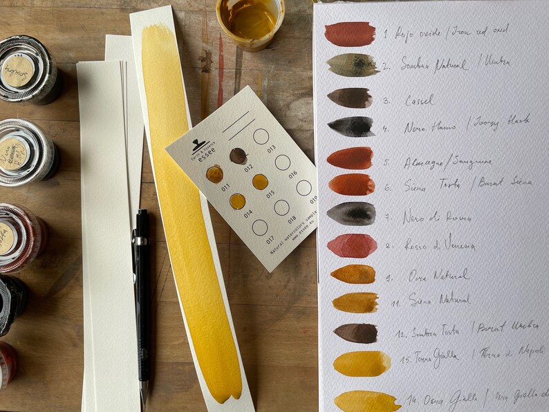 Puede incluir: Una carta de pintura acuarela con 14 tonos diferentes de marr&oacute;n, naranja y amarillo. La carta est&aacute; etiquetada con los nombres italiano e ingl&eacute;s de cada color. La carta est&aacute; en una hoja de papel blanco y est&aacute; rodeada de otros materiales de arte.
