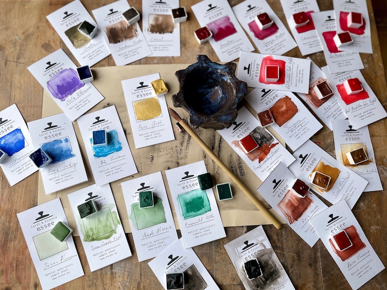 May include: An assortment of watercolour paint pans in various colours, including teal, green, red, gold, and black. The pans are arranged on a wooden surface with their corresponding labels, which feature the brand name "Earth & Essence essee" and the colour name.