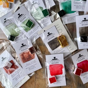 May include: An assortment of watercolour paint samples in various colours, including red, green, brown, and black. Each sample is labelled with the name of the colour and the brand "Earth & Essence essee".