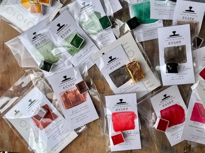 May include: An assortment of watercolour paint samples in various colours, including red, green, brown, and black. Each sample is labelled with the name of the colour and the brand "Earth & Essence essee".