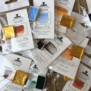 May include: A collection of small watercolour paint pans in various colours, including blue, orange, brown, and green. Each pan is labelled with the brand name "Earth & Essence essee" and the colour name. The pans are packaged in clear plastic bags with a white label.