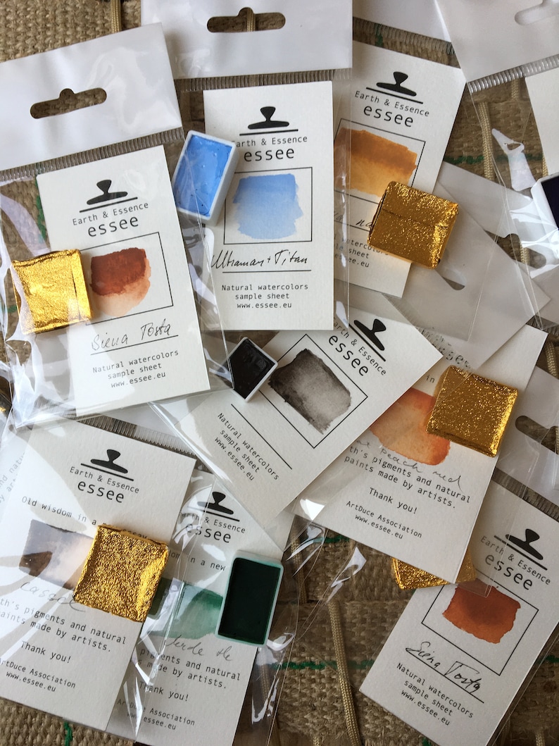 May include: A collection of small watercolour paint pans in various colours, including blue, orange, brown, and green. Each pan is labelled with the brand name "Earth & Essence essee" and the colour name. The pans are packaged in clear plastic bags with a white label.