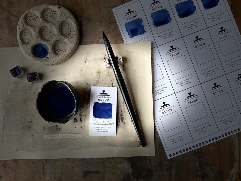 Op de afbeelding: Een klein kommetje met indigo blauwe aquarelverf, een penseel en een vel papier met kleurstalen voor Earth & Essence essee natuurlijke aquarelverf. De tekst op de kleurstaalkaart luidt "Indigo Blue Radix, Natuurlijke aquarelverf, Kleurstaal, E&eacute;n ounce per gewicht."
