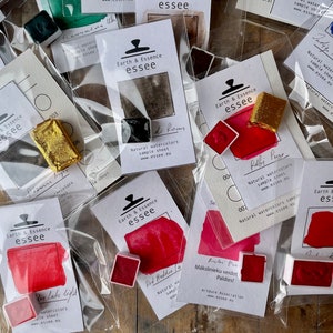 May include: An assortment of watercolour paint pans in various colours, including red, gold, and black. Each pan is labelled with the brand name "Earth & Essence essee" and a description of the colour. The pans are packaged in clear plastic bags.
