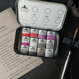 May include: A set of 12 artist-grade watercolors in a metal tin, with a variety of colors including red, pink, and gray. The tin is open, revealing the watercolor cakes, and is accompanied by a paintbrush and a card with color names.