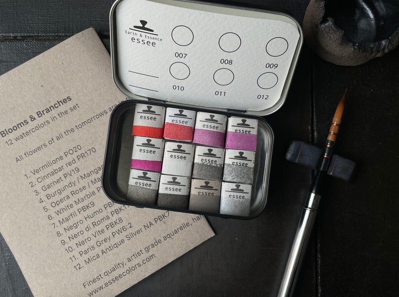May include: A set of 12 artist-grade watercolors in a metal tin, with a variety of colors including red, pink, and gray. The tin is open, revealing the watercolor cakes, and is accompanied by a paintbrush and a card with color names.