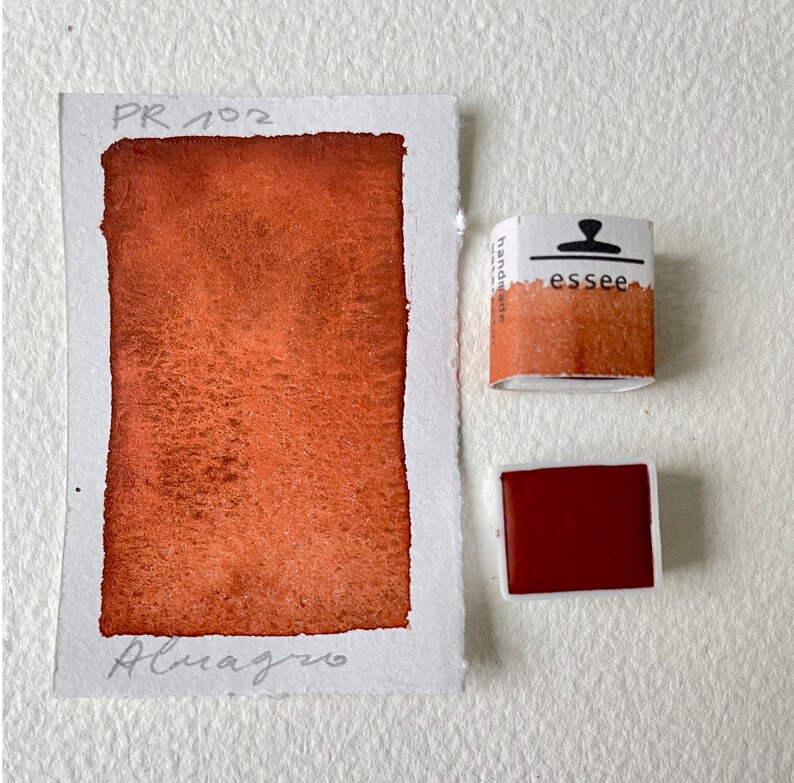 Puede incluir: Una paleta de pintura acuarela etiquetada como "PR 102" con el nombre "Almagro" escrito debajo. La pintura es de color rojo anaranjado oscuro. La paleta es de la marca "essee" y est&aacute; etiquetada como "handmade".