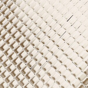 May include: An overhead shot of a repeating pattern of small, off-white, square-shaped containers. The containers are arranged in a grid-like pattern, creating a textured surface with shadows. The overall effect is a geometric, abstract design.
