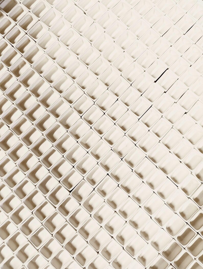 May include: An overhead shot of a repeating pattern of small, off-white, square-shaped containers. The containers are arranged in a grid-like pattern, creating a textured surface with shadows. The overall effect is a geometric, abstract design.