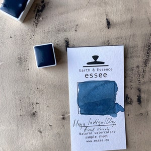 Op de afbeelding: Een monsterblad voor Maya Indigo/Clay Azul Verde natuurlijke aquarelverf van Earth & Essence essee. Het blad toont een blauwe aquarelkleurvlek en bevat het websiteadres van het bedrijf.