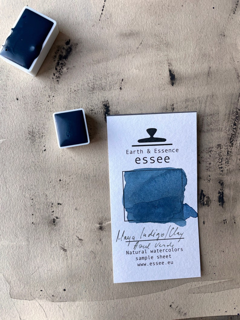 Op de afbeelding: Een monsterblad voor Maya Indigo/Clay Azul Verde natuurlijke aquarelverf van Earth & Essence essee. Het blad toont een blauwe aquarelkleurvlek en bevat het websiteadres van het bedrijf.