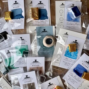 May include: A collection of watercolour paint samples in various colours, including blue, green, and gold. Each sample is labelled with the brand name "Earth & Essence essee" and a description of the colour. The samples are displayed on white cards with black text.