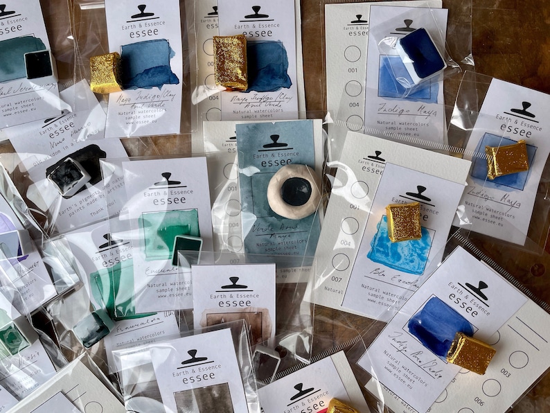 May include: A collection of watercolour paint samples in various colours, including blue, green, and gold. Each sample is labelled with the brand name "Earth & Essence essee" and a description of the colour. The samples are displayed on white cards with black text.