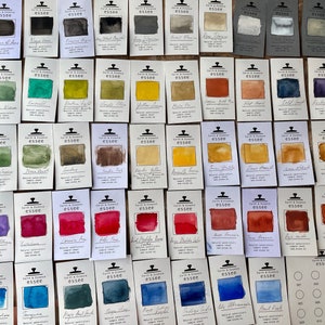Puede incluir: Una colecci&oacute;n de 60 muestras de pintura acuarela en varios colores, dispuestas en filas sobre una superficie blanca. Cada muestra est&aacute; etiquetada con el nombre del color y la marca "essee".