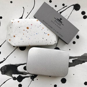 May include: A white rectangular object with colorful paint splatters, a silver rectangular tin, and a gray card with the text "essee Earth & Essence Ancient Wisdom in a new essay." The background has black ink splatters.
