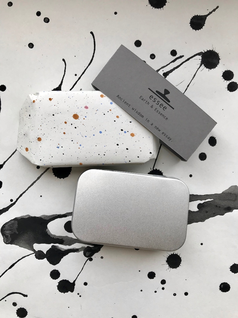 May include: A white rectangular object with colorful paint splatters, a silver rectangular tin, and a gray card with the text "essee Earth & Essence Ancient Wisdom in a new essay." The background has black ink splatters.