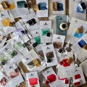 May include: An assortment of watercolour paint pans in various colours, including gold, green, blue, red, and brown. The pans are packaged in clear plastic bags with white labels that include the brand name "Earth & Essence essee" and the colour name.