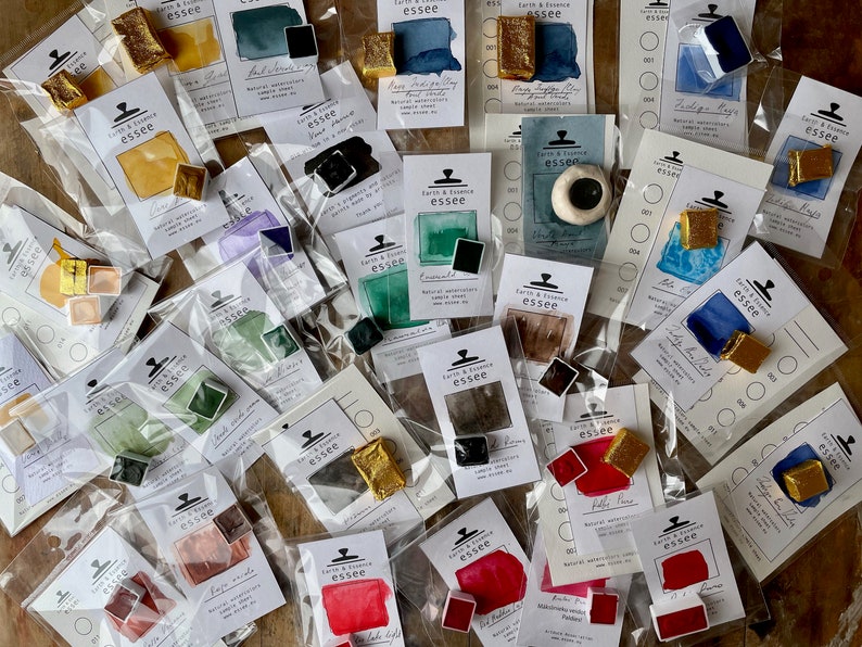May include: An assortment of watercolour paint pans in various colours, including gold, green, blue, red, and brown. The pans are packaged in clear plastic bags with white labels that include the brand name "Earth & Essence essee" and the colour name.