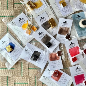 May include: An assortment of watercolour paint pans in various colours, including red, orange, yellow, brown, and black. The pans are labelled with the brand name "Earth & Essence essee" and the colour name. The pans are arranged on a hessian surface.
