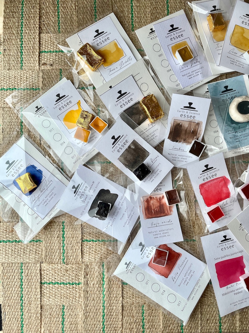 May include: An assortment of watercolour paint pans in various colours, including red, orange, yellow, brown, and black. The pans are labelled with the brand name "Earth & Essence essee" and the colour name. The pans are arranged on a hessian surface.