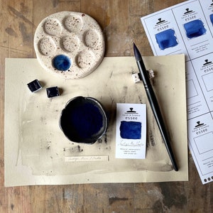 Op de afbeelding: Een klein keramisch palet met blauwe aquarelverf in een putje. Een klein kommetje met indigo-blauwe aquarelverf, twee aquarelverfpannetjes en een penseel zijn ook te zien. De afbeelding bevat een monsterkaart voor "Indigo Blue" aquarelverf van Earth & Essence Essee.