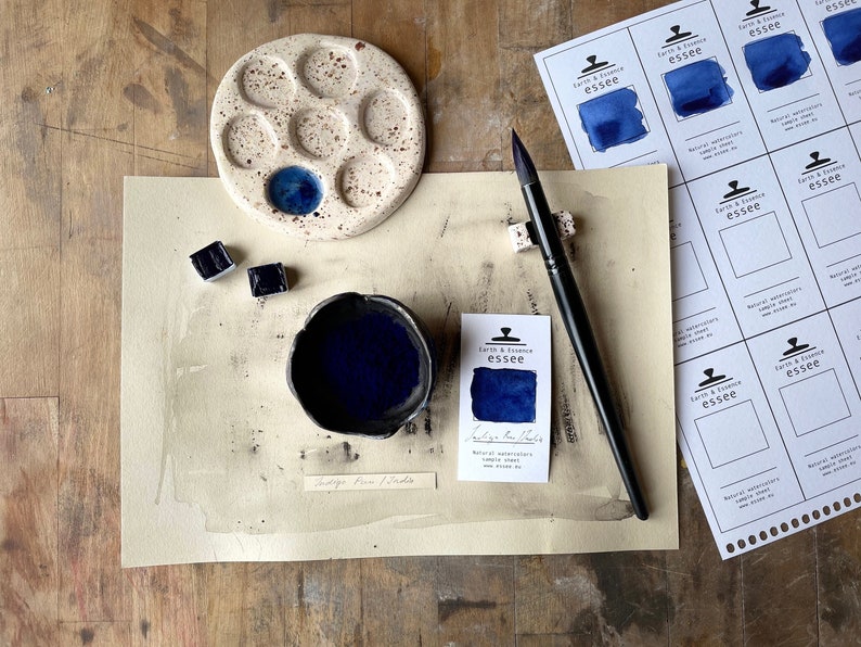 Op de afbeelding: Een klein keramisch palet met blauwe aquarelverf in een putje. Een klein kommetje met indigo-blauwe aquarelverf, twee aquarelverfpannetjes en een penseel zijn ook te zien. De afbeelding bevat een monsterkaart voor "Indigo Blue" aquarelverf van Earth & Essence Essee.